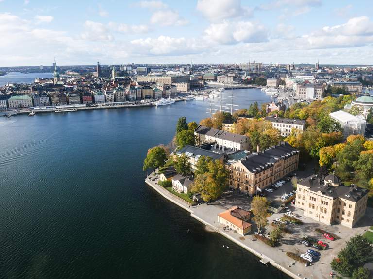 En helikopterbild över Stockholms innerstad på hösten, med Skeppsholmens träd klädda i gula och orangea nyanser, samt Gamla Stans historiska byggnader i bakgrunden