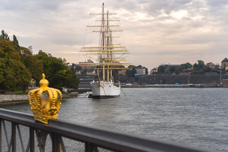 I förgrunden syns den stålgrå Skeppholmsbrons räcke, prydd med en gyllene krona. I bakgrunden syns det ståtliga vita örlogsskeppet Af Chapman, som ligger vid den gröna ön Skeppsholmens västra strand.