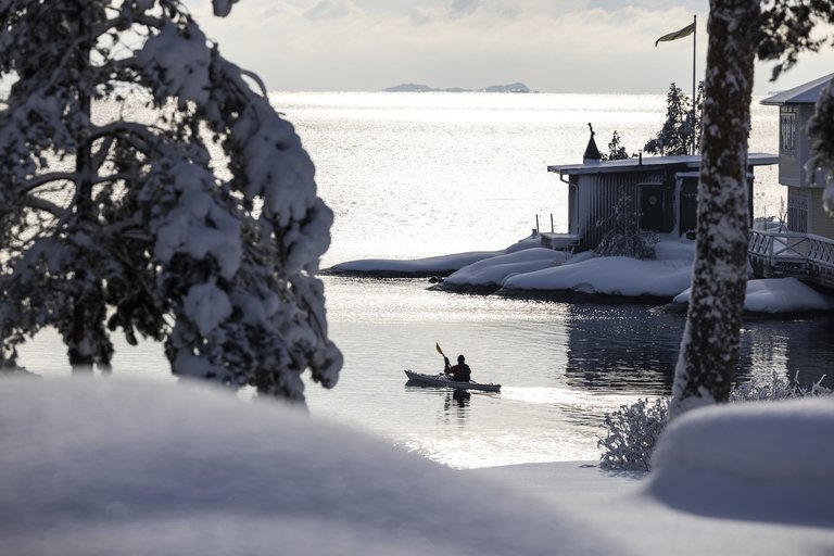 En kajak på lugnt vatten i motljus, omgiven av snöiga klippor och snötäckta tallar i Stockholms Skärgård