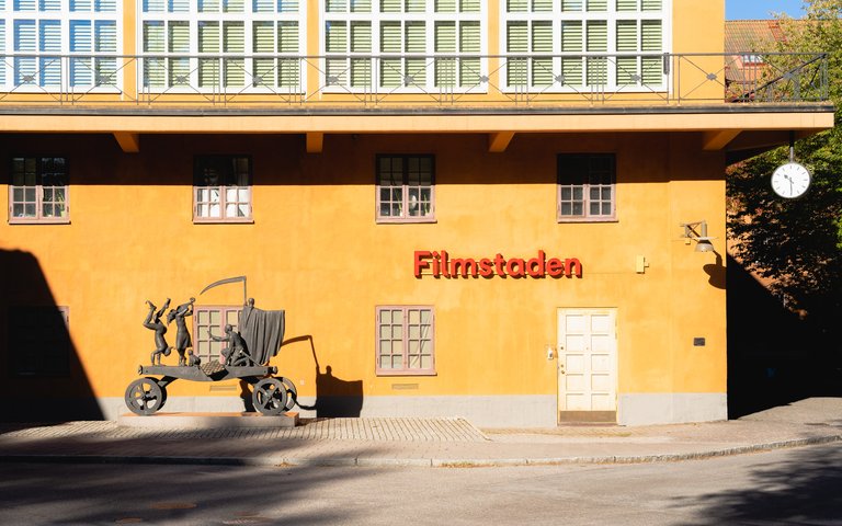 En staty står framför ett orange hus. Statyn föreställer Ingmar Bergman och en vagn full av gycklare.