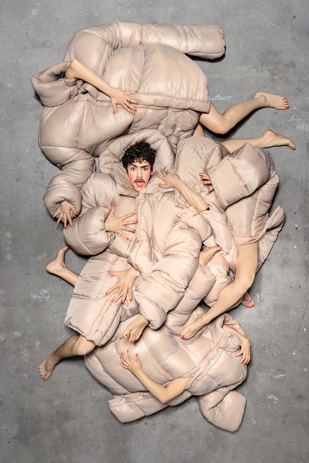 En person ligger på ett betonggolv omgiven av flera kroppar insvepta i beige, vadderade täcken. Armar, ben och händer sticker fram i olika riktningar och skapar en sammanflätad, skulptural formation. Personen i mitten har ett uttrycksfullt ansikte med öppen mun och blicken riktad uppåt.